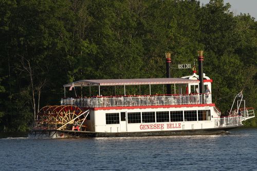 The image is of the Genesee Belle Paddleboat at the crossroads of the village. It is a two-decker boat floating down the river.