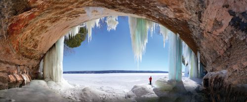 The image is of a brown crave with ice casting over the entrance. Outside this cave, there is a white sheet of ice-over Lake Superior. Far in the distance there stands a man in a red coat. There is a crystal clear blue sky ahead. 