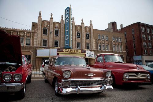 The image of the back of the bricks Capital Theatre. In front of the theater is a group of antique cars. 