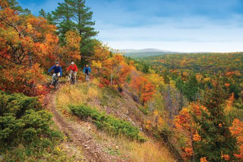The image is of Copper Harbor in Michigan. It's a beautiful fall day on a trail in the hills. The trees are various colors of orange, light green, dark green, and red. On the top left of this trail are three bikers with helmets riding down the hill.